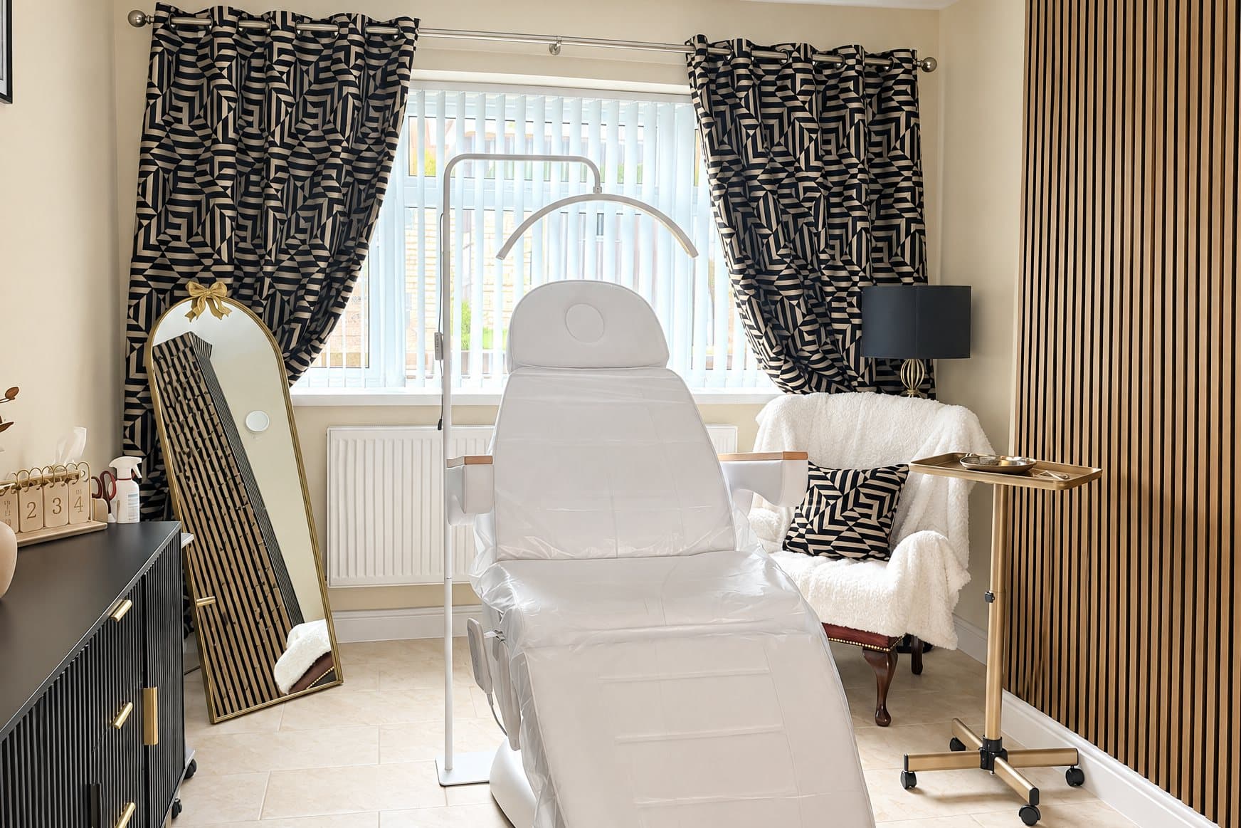 A white reclining treatment chair under a window with black-and-cream geometric curtains, between a gold arched floor mirror and a white armchair, with a gold tray trolley beside a gold slatted wall